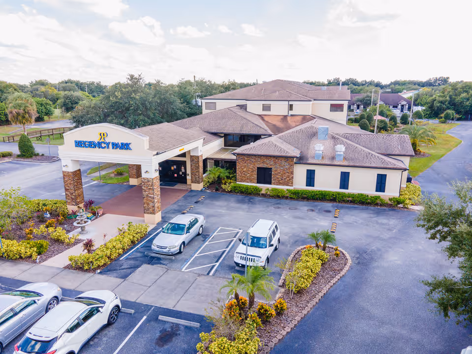 Aerial view of Regency Park Assisted Living & Memory Care facility showing the main building with a covered entrance, parking lot with several cars, landscaped garden areas with shrubs and small palm trees, and surrounding greenery under a partly cloudy sky.