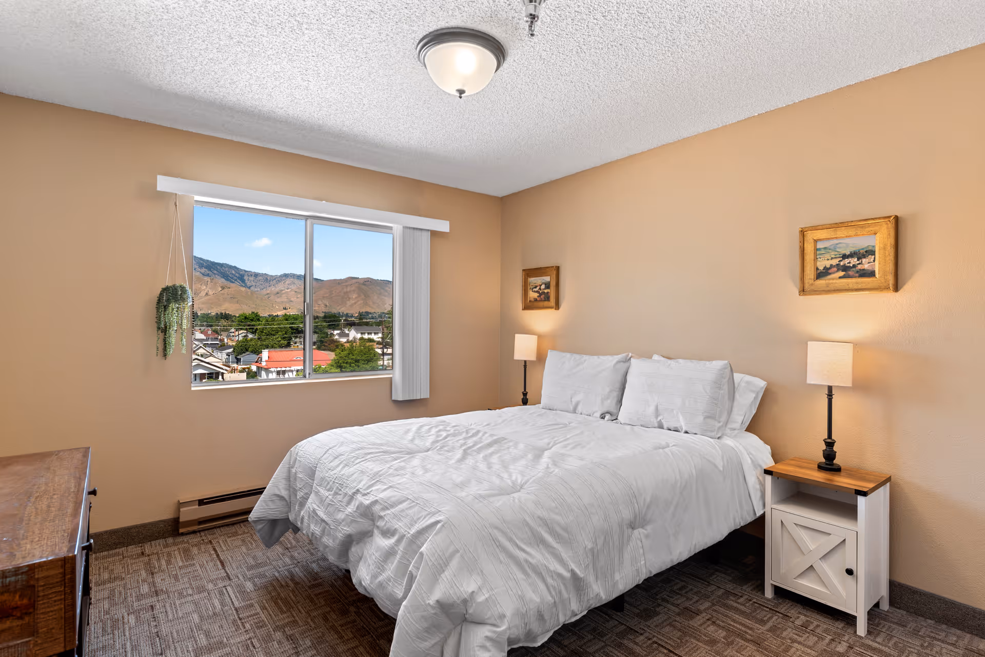 A cozy bedroom with beige walls and a large window showing a view of mountains and houses outside. The room features a neatly made bed with white bedding and pillows, two bedside tables each with a lamp, a wooden dresser, two small framed paintings on the walls, and a hanging plant near the window.