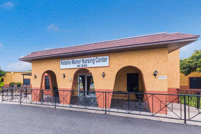 Exterior view of a single-story building with a sign that reads 'Retama Manor Nursing Center 636'. The building has a yellow and reddish-brown stucco finish with three archways at the front entrance and black metal railings along the sidewalk. There is a clear blue sky in the background.
