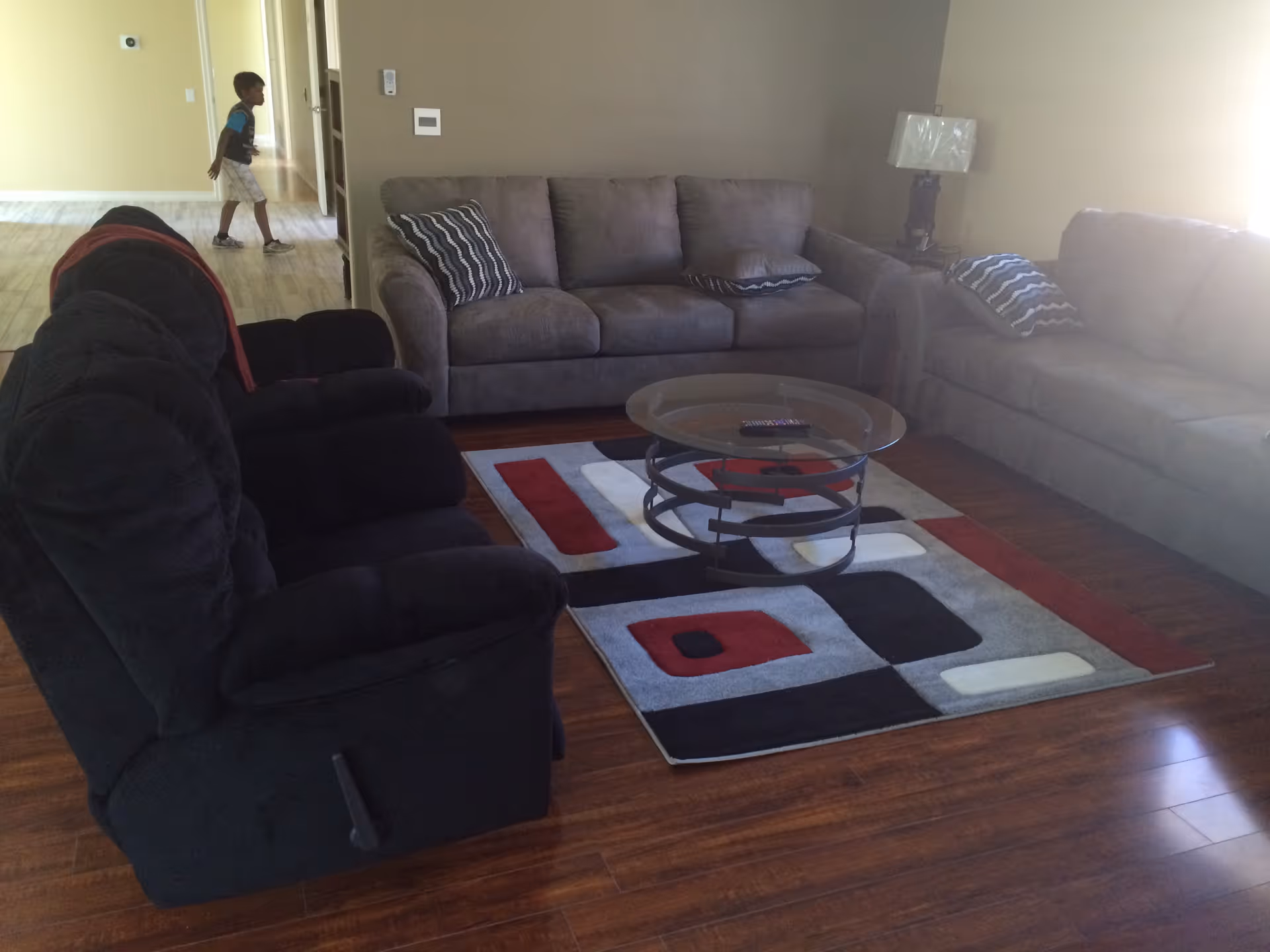 A living room with two gray sofas and a black recliner chair arranged around a glass coffee table on a patterned rug. A lamp is on a side table next to one sofa. In the background, a child is walking in a hallway with light-colored flooring.