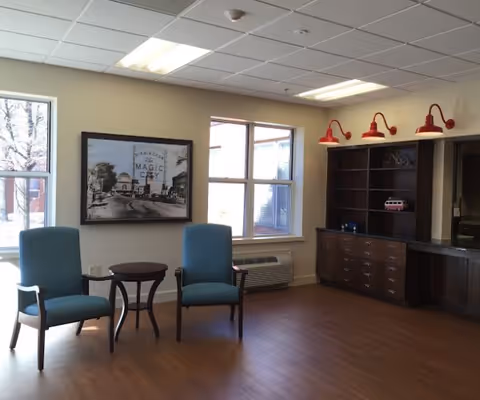 A room with two blue armchairs and a small round wooden table between them. The room has wooden flooring, a large window letting in natural light, a framed black and white photo on the wall, and a dark wooden cabinet with red wall-mounted lamps above it.