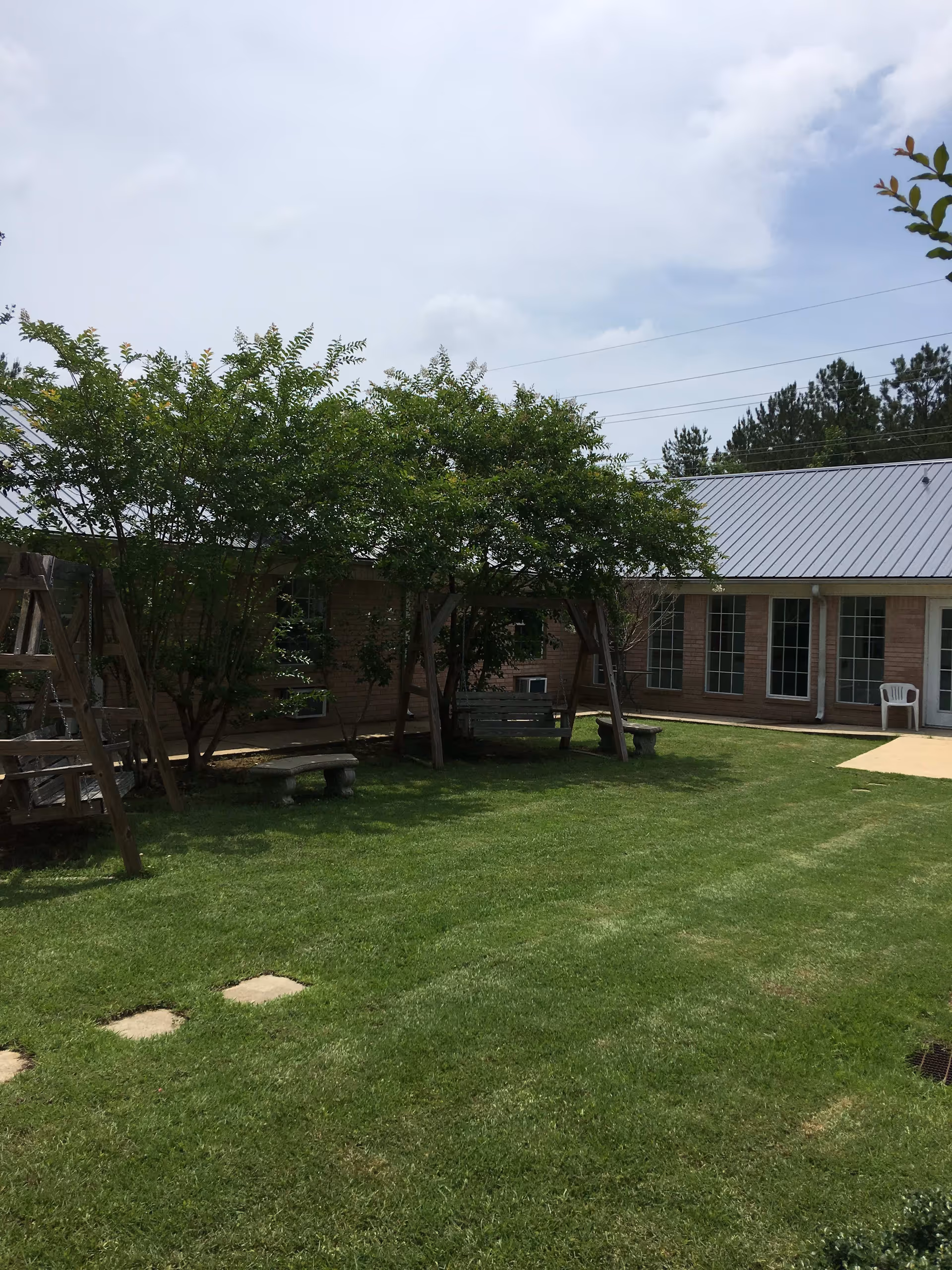 A grassy outdoor courtyard area with two wooden swings and stone benches under leafy trees. The courtyard is surrounded by a single-story brick building with multiple windows and a metal roof. A white plastic chair is visible near a door on the right side.