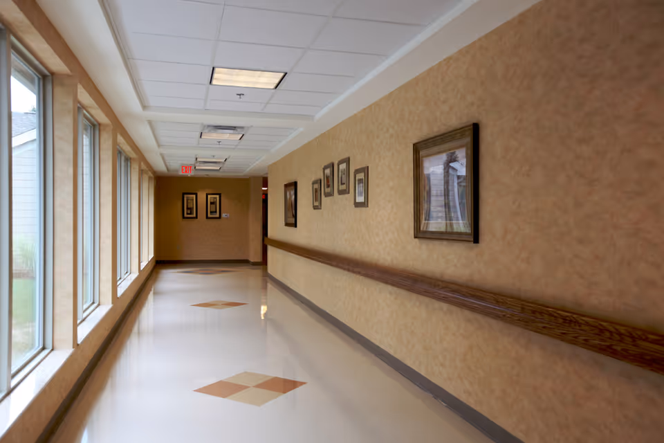 A clean, well-lit hallway in a senior living facility with large windows on the left side letting in natural light. The right wall is decorated with framed pictures and has a wooden handrail running along its length. The floor is polished with diamond-shaped tile patterns, and the ceiling has recessed lighting panels.
