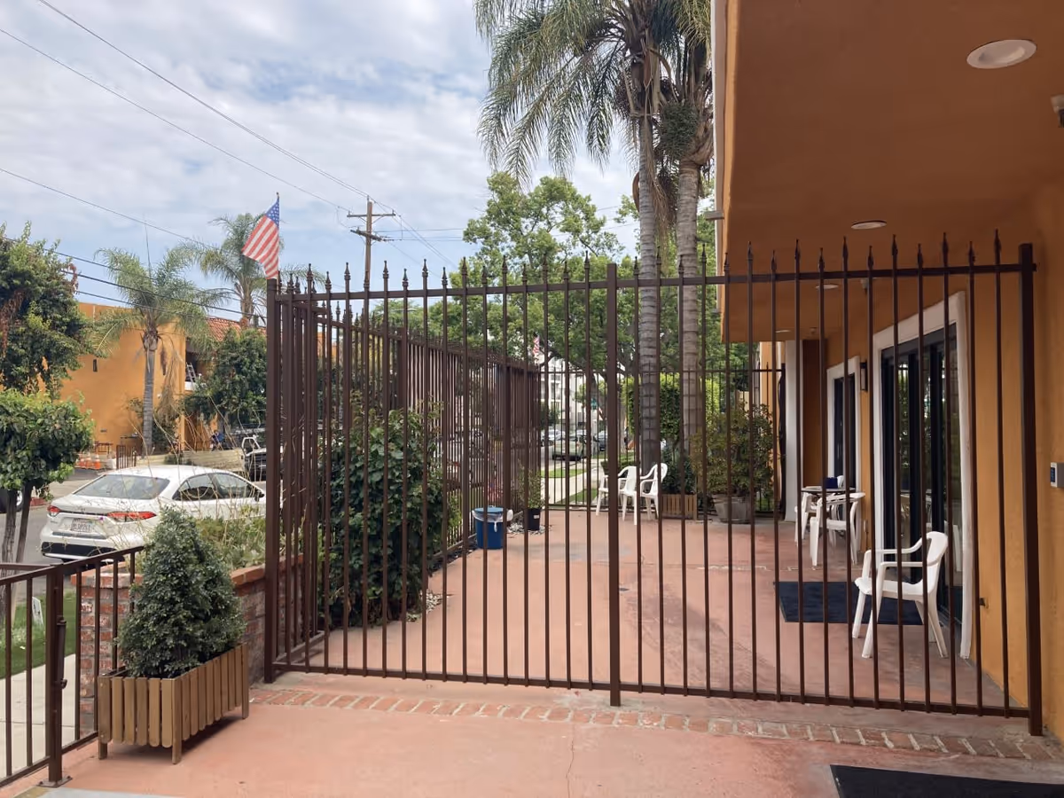 Outdoor patio area of a senior living facility with a brown metal fence gate in the foreground. The patio has several white plastic chairs and potted plants. The building exterior is painted in a warm tan color with large sliding glass doors. Palm trees and other greenery are visible, along with a parked white car and an American flag in the background under a partly cloudy sky.