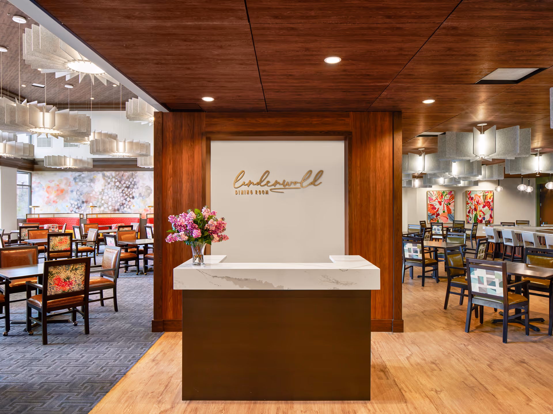 Interior view of a dining room with a central reception desk featuring a vase of pink and purple flowers. The wall behind the desk has a sign that reads 'Underwell Dining Room'. The room has wooden ceilings, modern light fixtures, and multiple tables and chairs arranged for dining. Colorful artwork is visible on the walls in the background.
