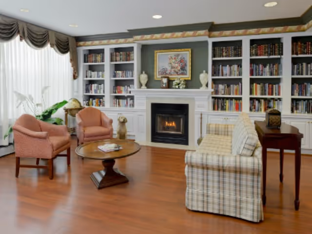 A cozy living room with a lit fireplace surrounded by built-in bookshelves filled with books. The room features a plaid sofa, two upholstered armchairs, a round wooden coffee table, and a side table. Large windows with sheer curtains and valances allow natural light to fill the space. Decorative items including a globe, plants, and framed artwork are also visible.