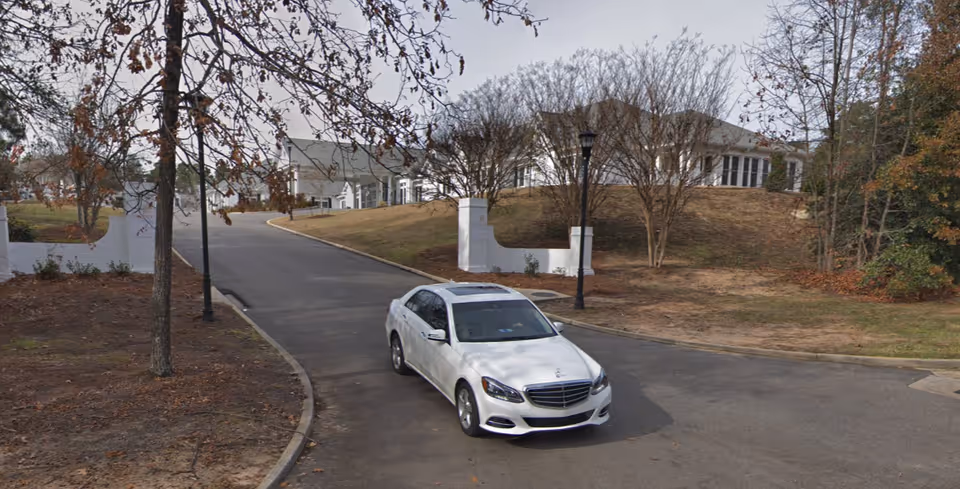A white sedan on a curved driveway leading to the entrance and pillars of a white, hilltop senior living building with trees and lamp posts.