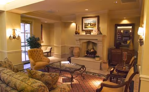 A cozy living room with a stone fireplace, a glass coffee table, a floral patterned sofa, a yellow armchair, and wooden chairs. The room is warmly lit with wall sconces and recessed ceiling lights. There is a framed painting above the fireplace and decorative vases on the mantel. A large mirror and a wooden cabinet are visible on the right side of the room.