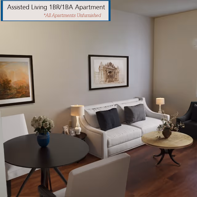 A furnished living room in an assisted living 1 bedroom, 1 bathroom apartment featuring a white sofa with two dark cushions, a round coffee table with a flower vase, a round dining table with white chairs and a flower vase, two table lamps, and framed artwork on the walls.