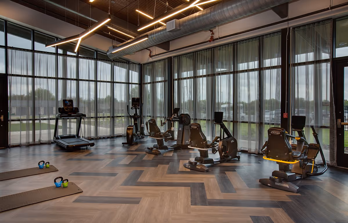 A modern fitness room with large floor-to-ceiling windows covered by sheer curtains. The room features various exercise equipment including treadmills, stationary bikes, and kettlebells placed on exercise mats. The ceiling has exposed ductwork and contemporary linear lighting fixtures.