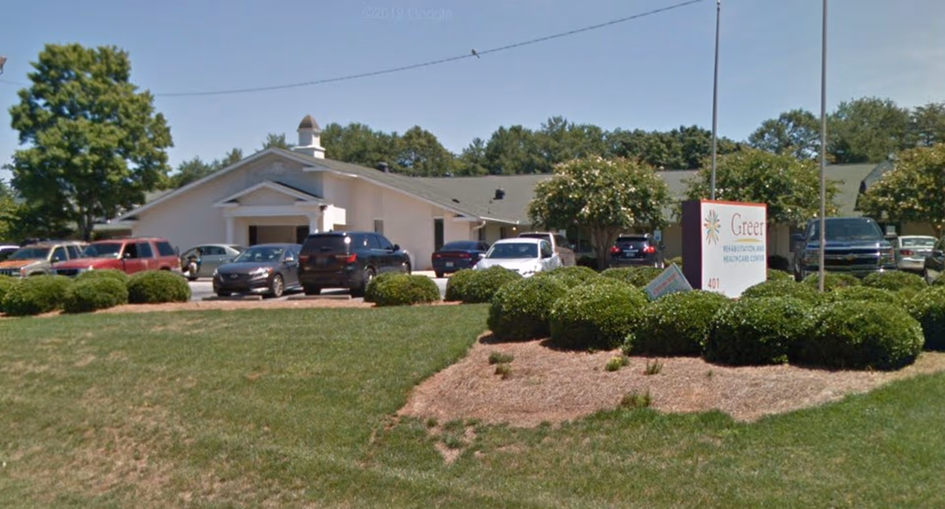 Exterior view of a single-story healthcare facility with a parking lot in front containing several cars. The building is surrounded by green bushes and trees under a clear blue sky. A sign in front reads 'Greer Rehabilitation and Healthcare Center.'