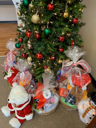 A decorated Christmas tree with red, green, and gold ornaments standing on a carpeted floor. In front of the tree are several gift baskets wrapped in clear plastic with colorful ribbons, and a white teddy bear wearing a red Santa outfit is sitting on the floor to the left.