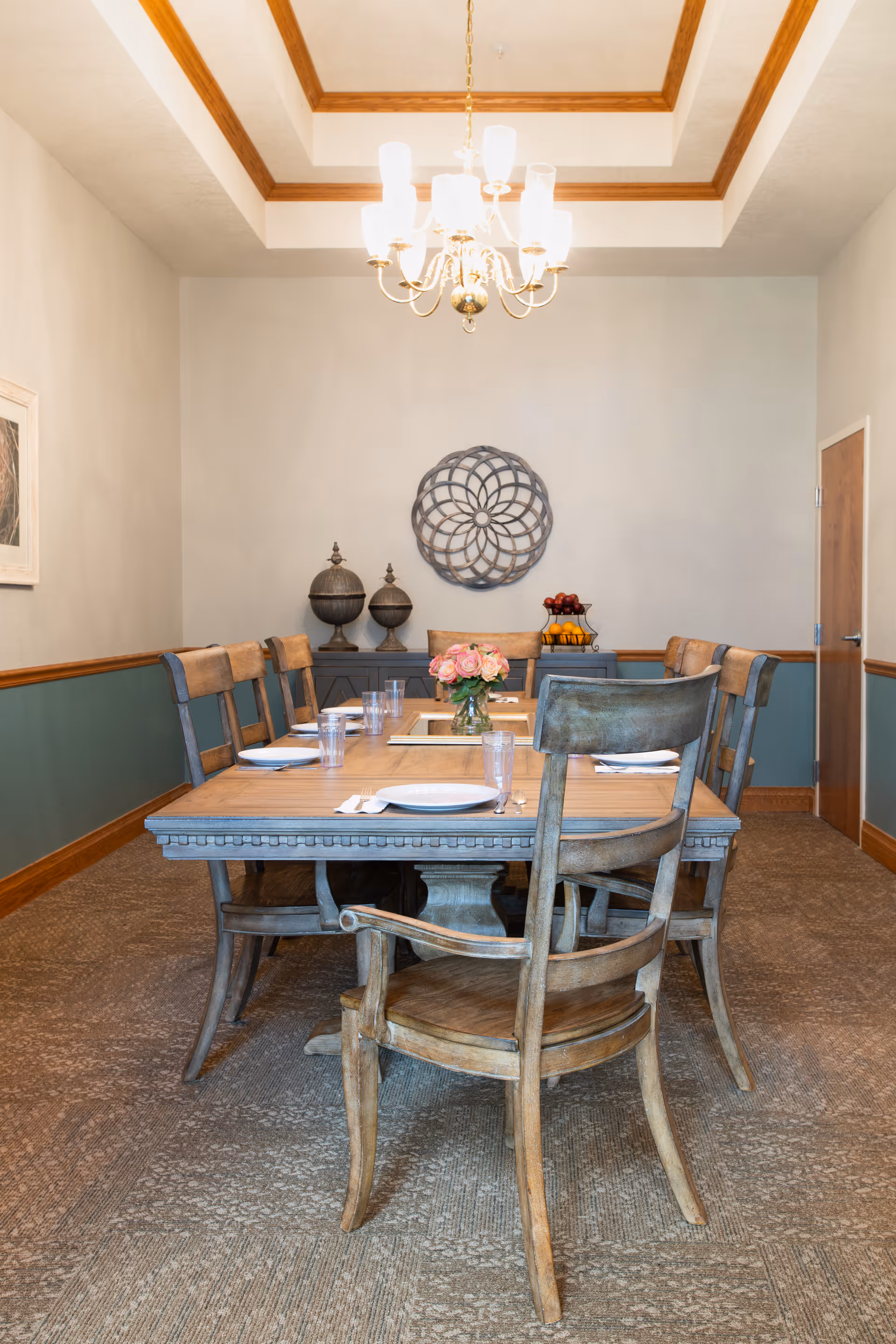 A formal dining room with a long wooden table set with plates and glasses, wooden chairs, a chandelier, and wall decor.