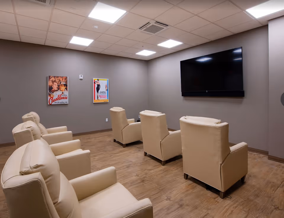 A small media room with six beige armchairs arranged in two rows facing a large flat-screen TV mounted on a gray wall. The room has wood flooring, a drop ceiling with recessed lighting, and two classic movie posters on the wall.