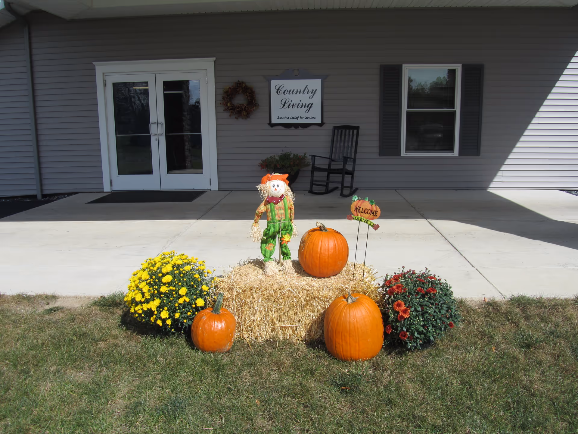 Entrance of a Country Living adult foster care building decorated with pumpkins, a scarecrow on a hay bale and potted mums.