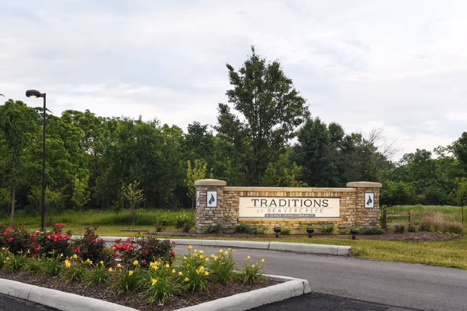 Stone entrance sign reading "Traditions of Beavercreek" surrounded by landscaping and trees.