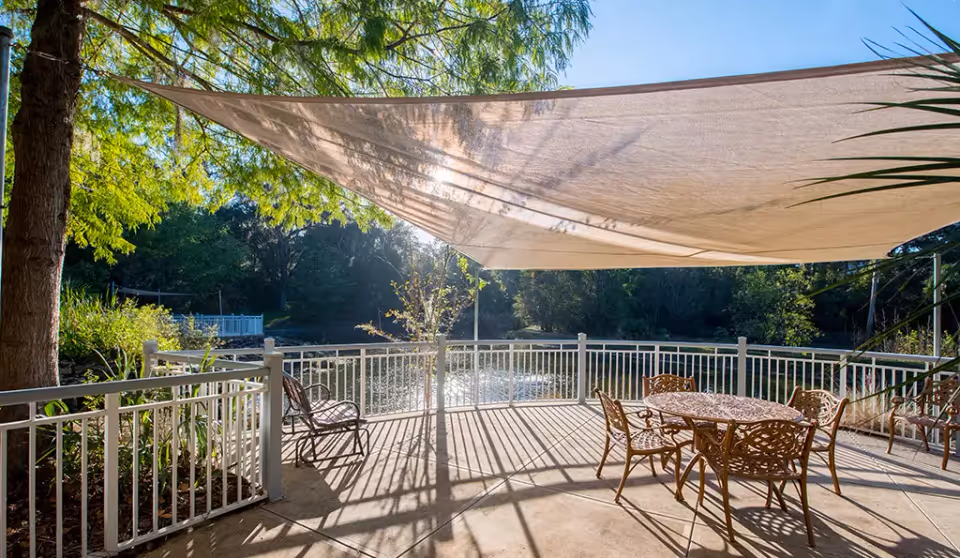 Shaded outdoor patio with metal table and chairs overlooking a pond surrounded by trees.
