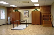 Physical therapy/rehab room with parallel walking bars on a mat, practice stairs, two wooden cabinets, and exercise equipment against a brick-accent wall.