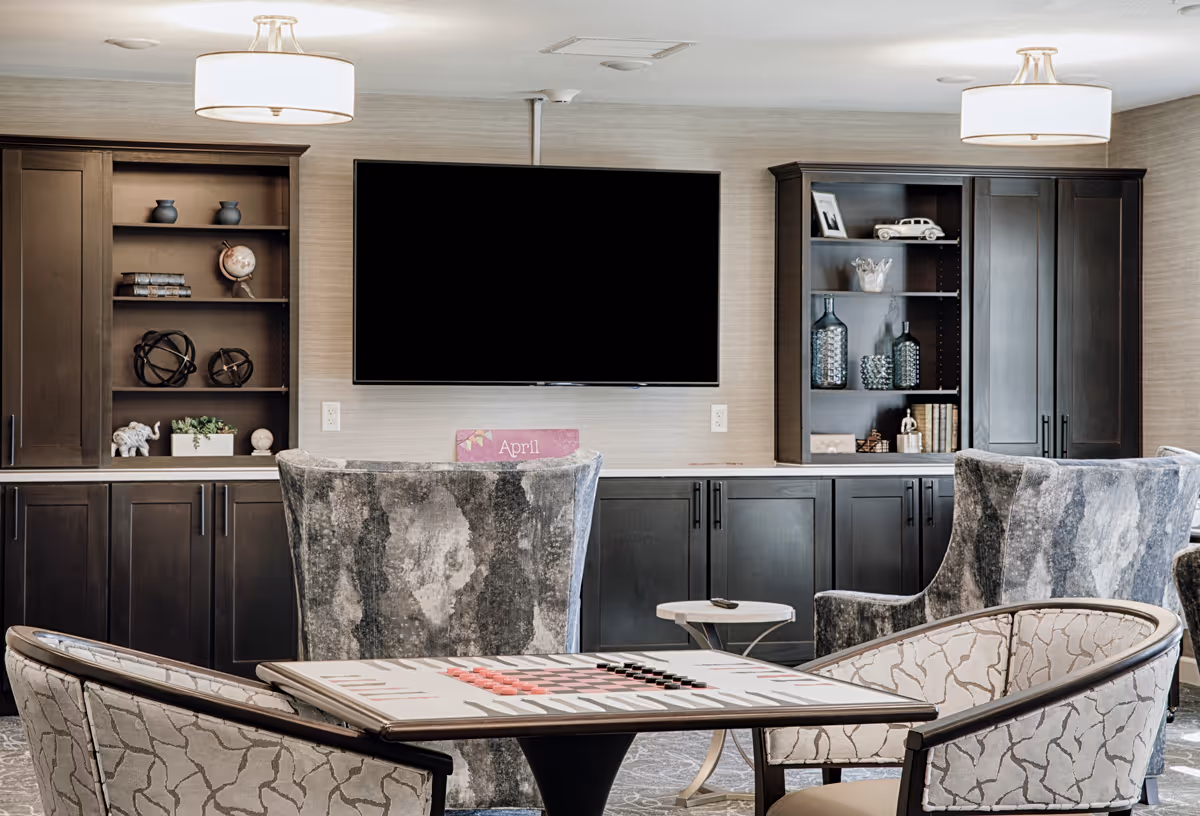 Communal living room with upholstered chairs around a game table, a wall-mounted TV, and built-in dark cabinets and shelving.