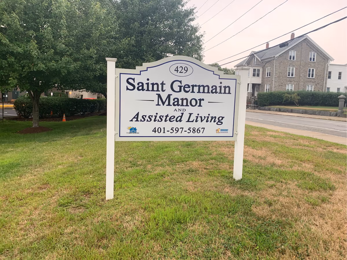 A white sign with blue trim on a grassy area near a sidewalk and street. The sign reads '429 Saint Germain Manor and Assisted Living 401-597-5867'. Trees and a multi-story stone and white building are visible in the background.