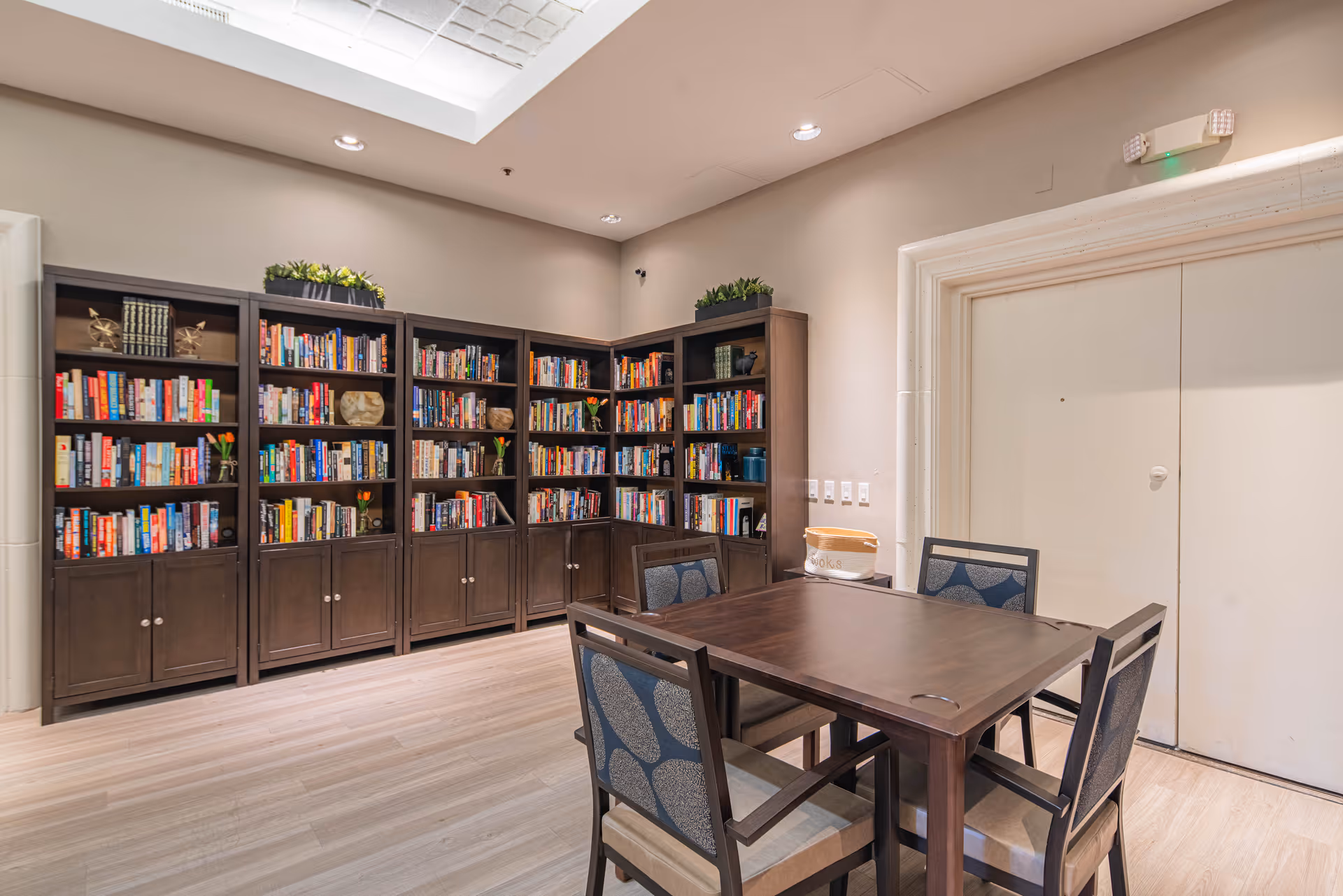 Well-lit communal library room with dark wooden bookshelves filled with books and a central table surrounded by chairs.