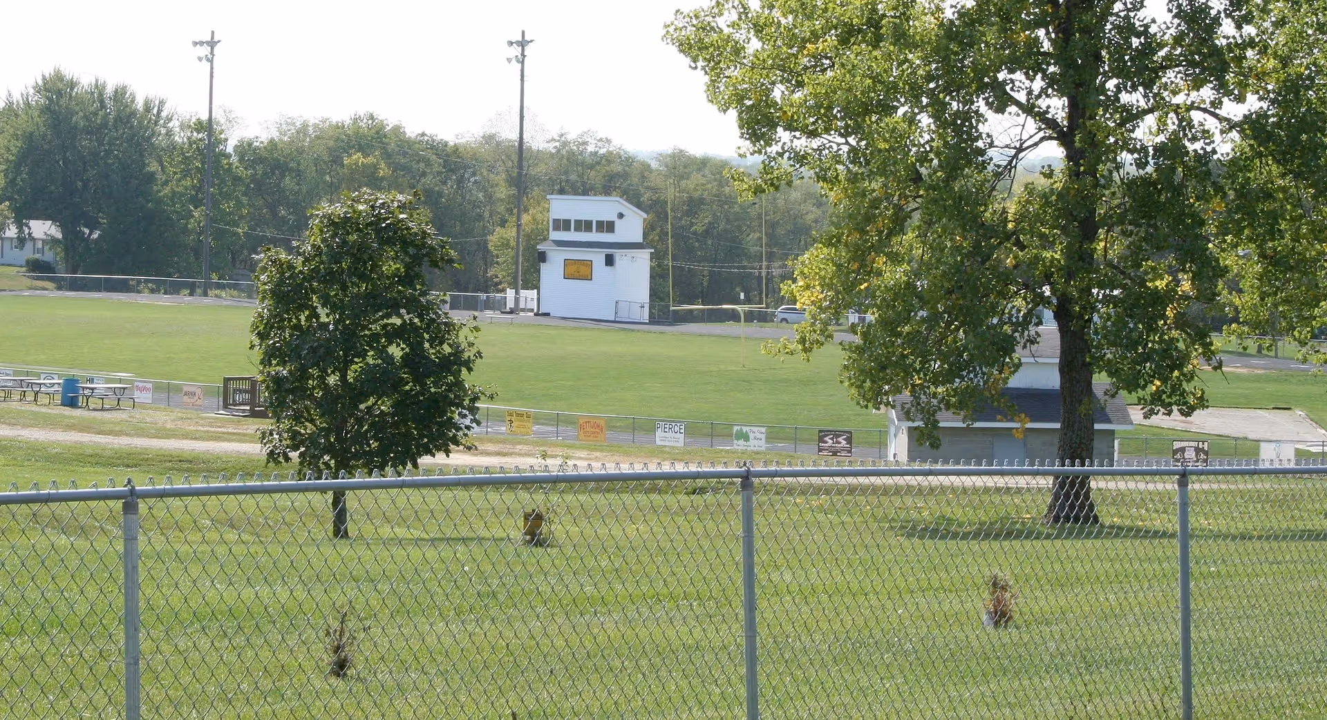 A fenced grassy field with two trees in the foreground and a sports field with a small white building and bleachers in the background under a clear sky.