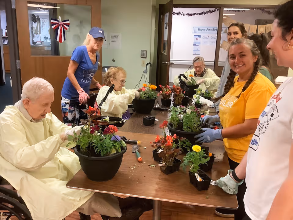 Several elderly individuals and caregivers are gathered around a table indoors, engaging in a flower planting activity. The elderly participants are seated and wearing protective gowns, while the caregivers stand nearby, smiling and assisting. Various potted flowers and gardening tools are on the table. The room has doors and bulletin boards in the background.