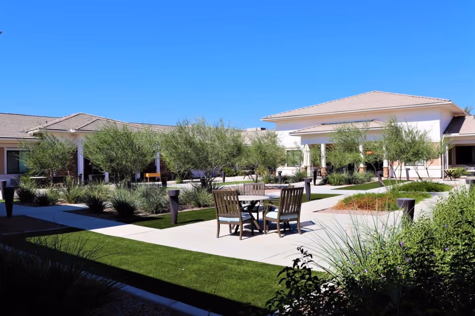 Outdoor courtyard area at Savanna House featuring a paved walkway, green grass patches, small trees, and a round table with four wooden chairs under a clear blue sky.
