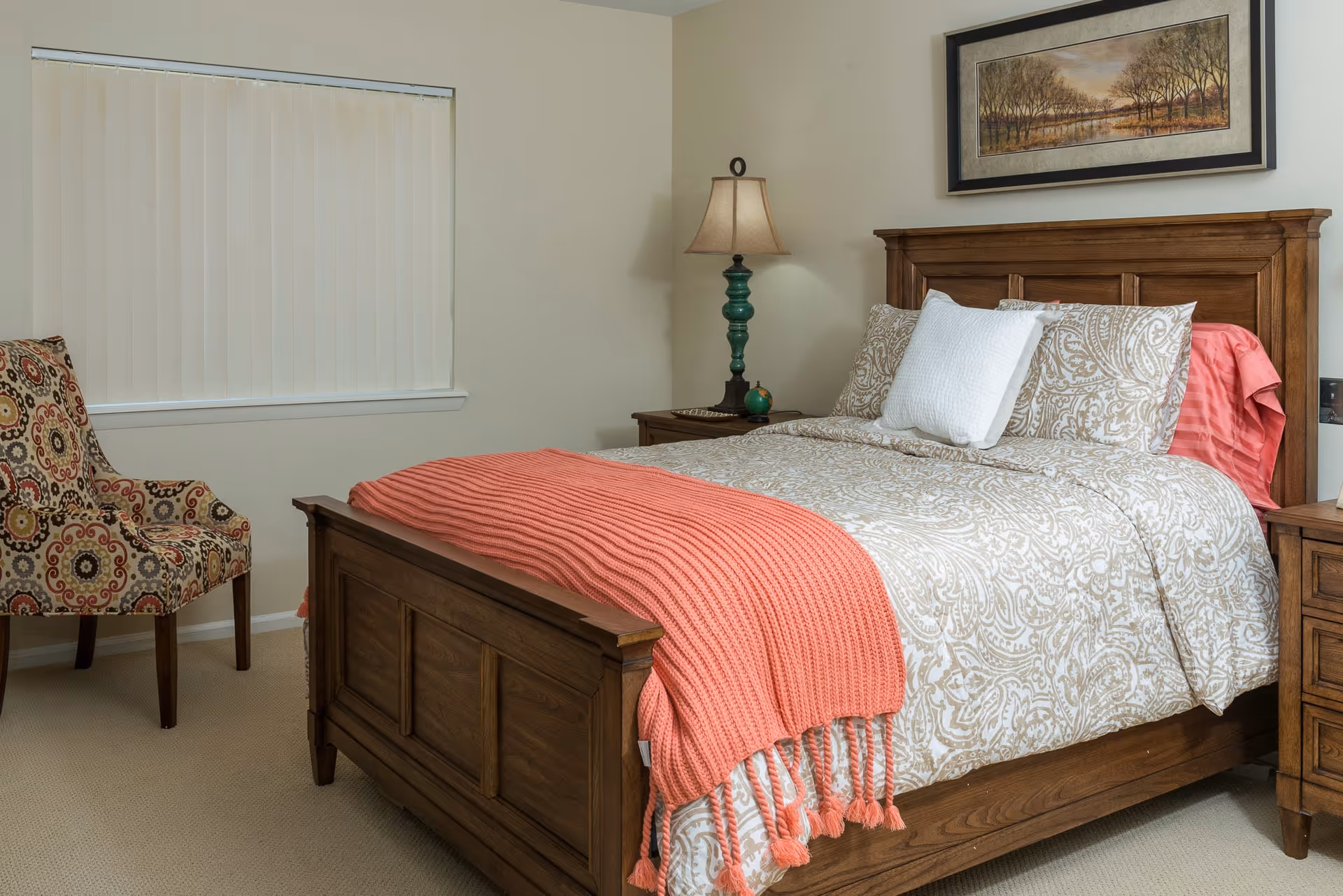 A cozy bedroom with a wooden bed frame, beige patterned bedding, a coral knitted throw blanket, a white pillow, a wooden nightstand with a green lamp, a framed landscape painting on the wall, a patterned armchair, and a window with vertical blinds.