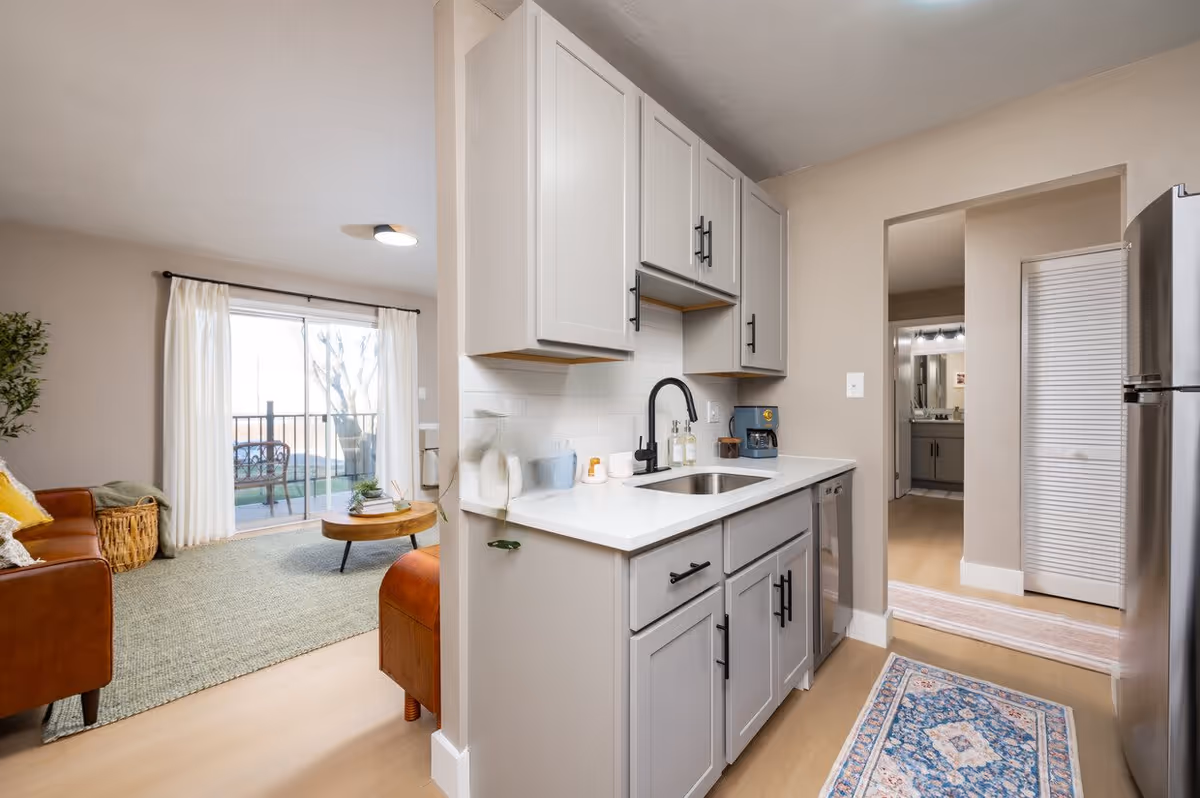 A modern kitchen featuring gray cabinetry, a stainless steel refrigerator, and a view of a living area with a balcony. Natural light floods the space through large windows, highlighting a cozy atmosphere.