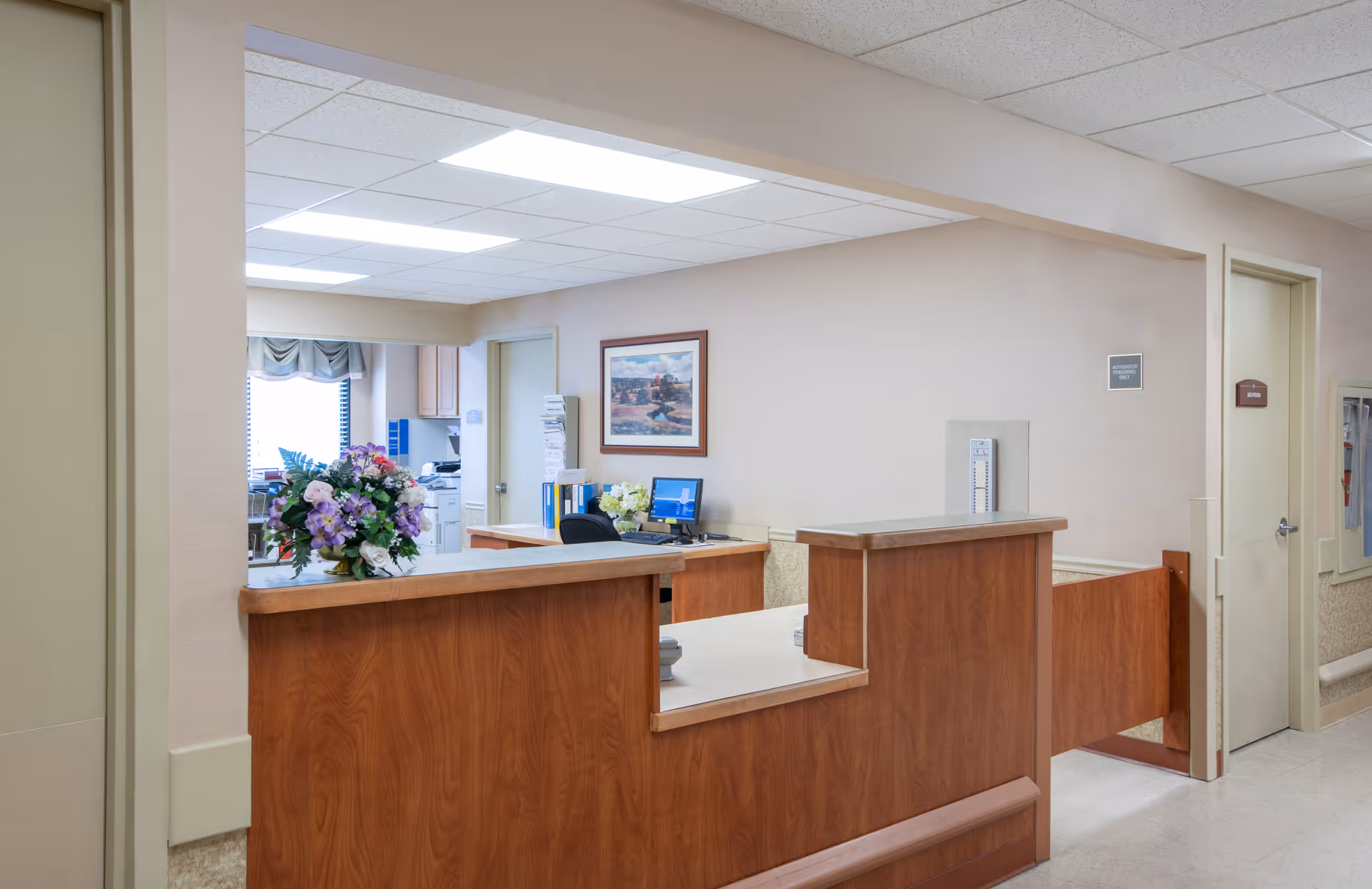 Reception desk area in a healthcare facility with a wooden counter, a computer, office chair, and a bouquet of flowers on the counter. The background shows a window with blinds, a framed picture on the wall, and a door leading to another room.