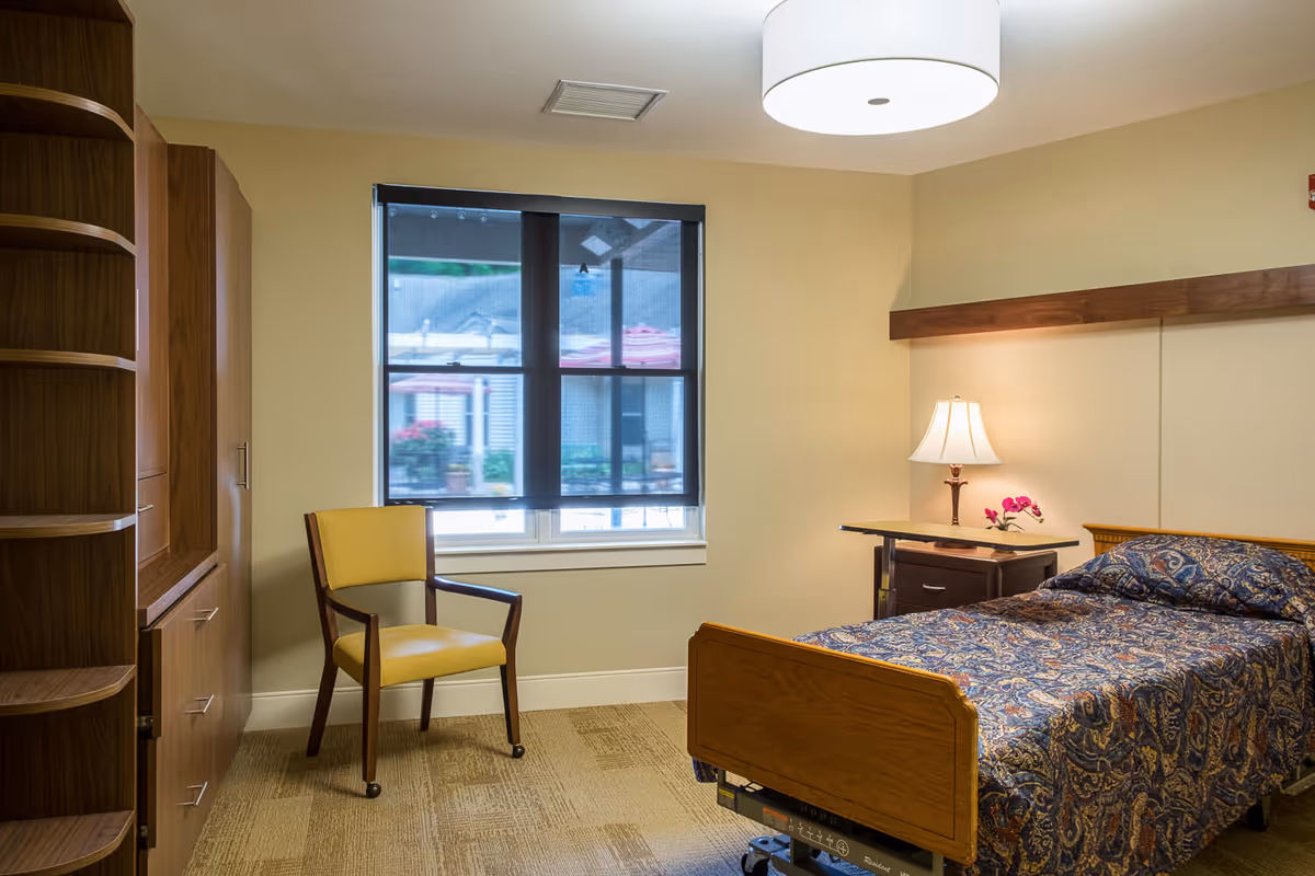 Well-lit private bedroom in a senior living facility with a single bed, bedside table and lamp, a yellow chair, and wooden cabinetry.
