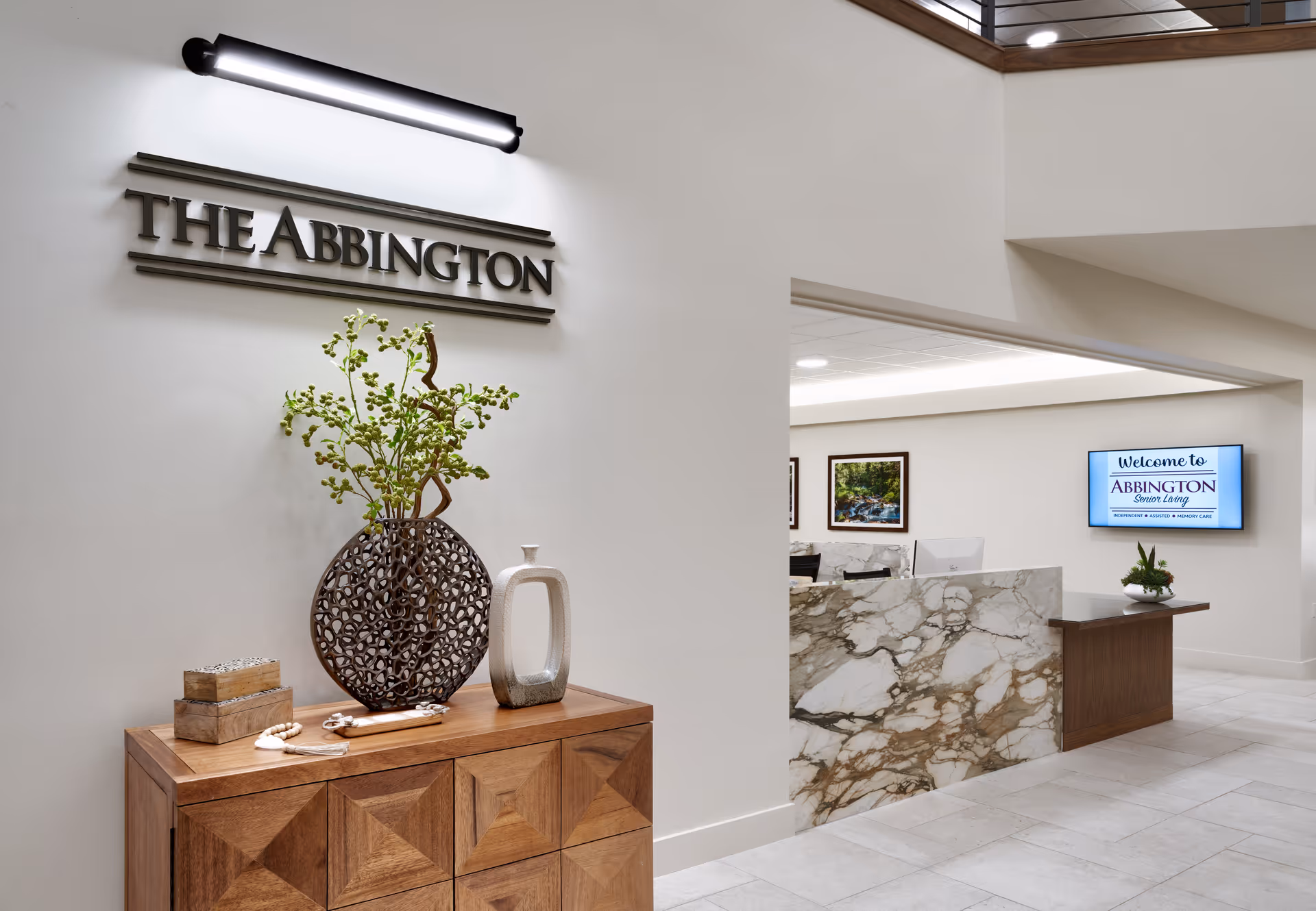 Reception area of The Abbington Senior Living facility featuring a wooden cabinet with decorative vases and a plant, a marble reception desk, and a wall-mounted screen displaying a welcome message.
