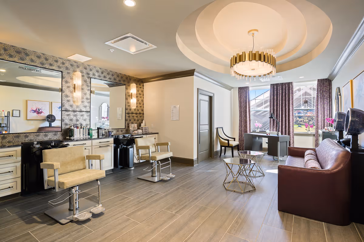 A bright and modern salon area in a senior living facility featuring two beige salon chairs in front of large mirrors with hair care products on the counter. The room has a stylish chandelier, a brown leather couch, a desk with a chair near a window with purple curtains, and decorative wall art.