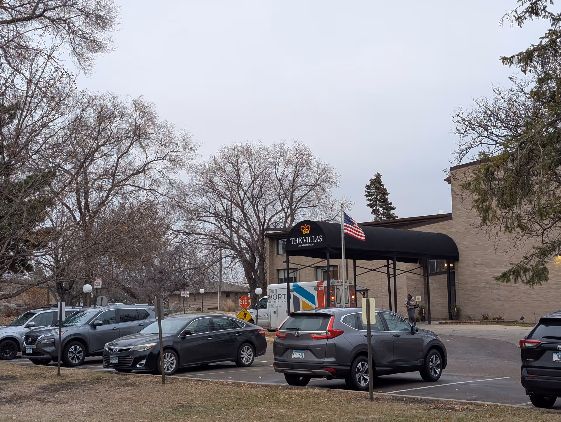 Exterior view of The Villas at Brookview building with a black awning displaying the facility name. Several cars are parked in front, and an American flag is flying near the entrance. Leafless trees and a cloudy sky are visible in the background.