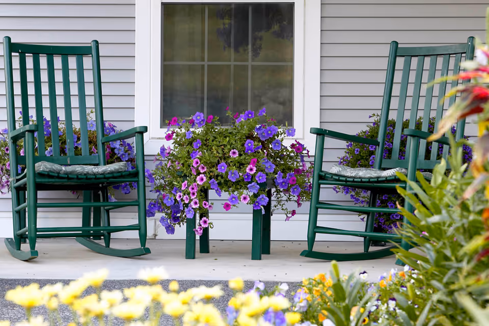 Two green wooden rocking chairs with cushions on a porch in front of a window, flanked by vibrant purple and pink flowers in pots, with additional colorful flowers in the foreground.