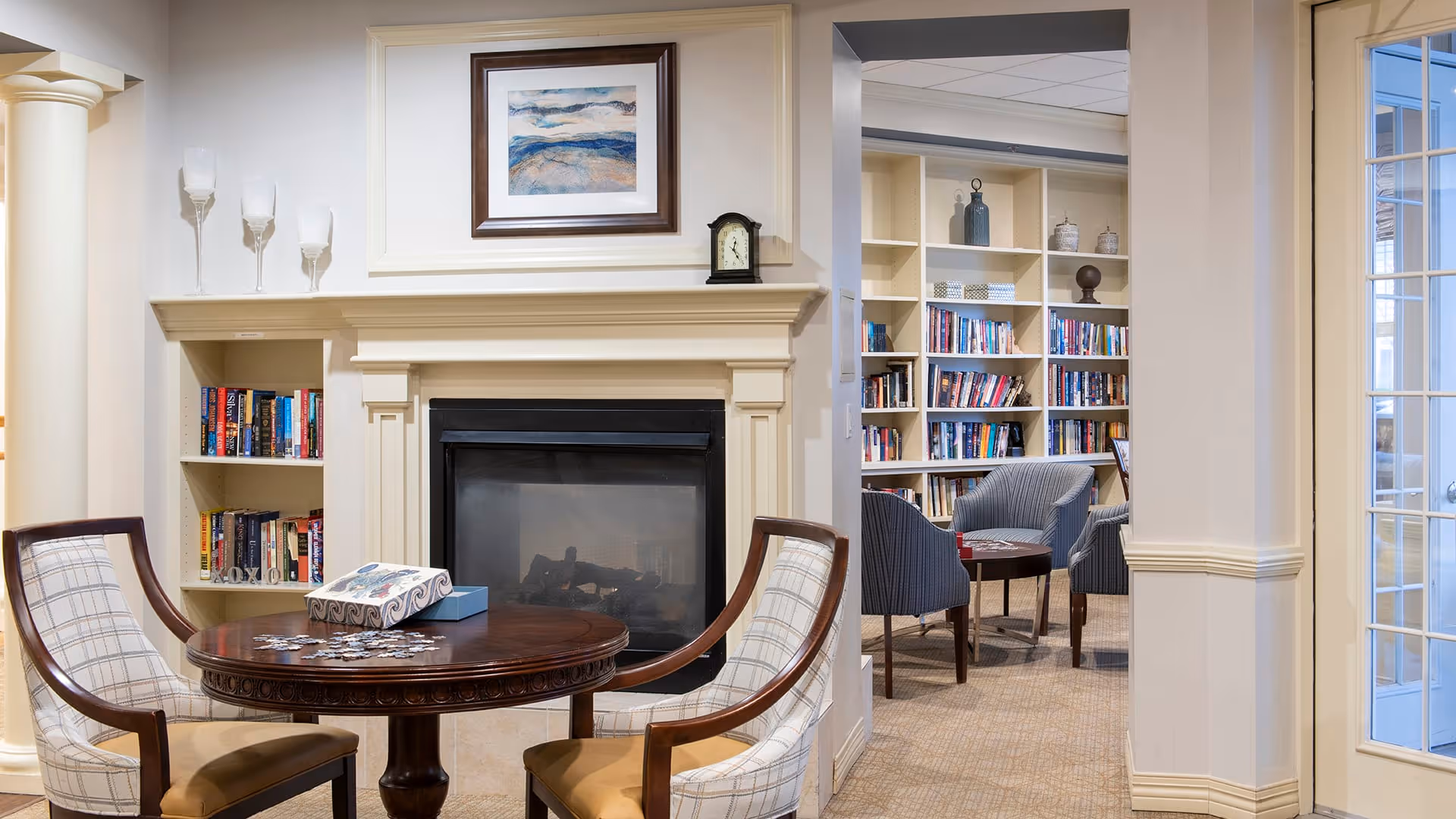Cozy interior common area with a round wooden table and two upholstered chairs in front of a fireplace. A partially completed puzzle and puzzle box are on the table. Built-in bookshelves filled with books flank the fireplace and extend into an adjacent room with additional seating and more bookshelves. Light-colored walls and carpeted floors create a warm and inviting atmosphere.