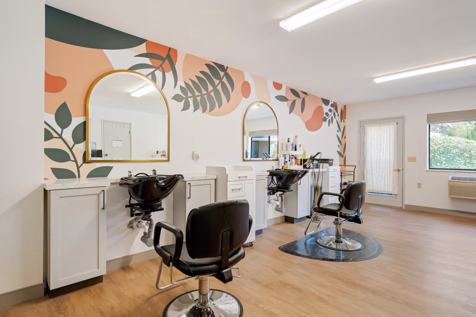 Interior view of a hair salon area in a retirement community with two black salon chairs in front of two washbasins and large mirrors mounted on a wall decorated with a colorful leaf and abstract pattern. The room has wood flooring, a door with a lace curtain, a window with a view of greenery outside, and bright overhead lighting.