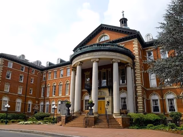 Exterior view of a large, historic brick building with tall white columns at the entrance and a curved portico. The building has multiple windows and a cupola on the roof. There are shrubs and trees around the building and a paved walkway leading to the entrance.