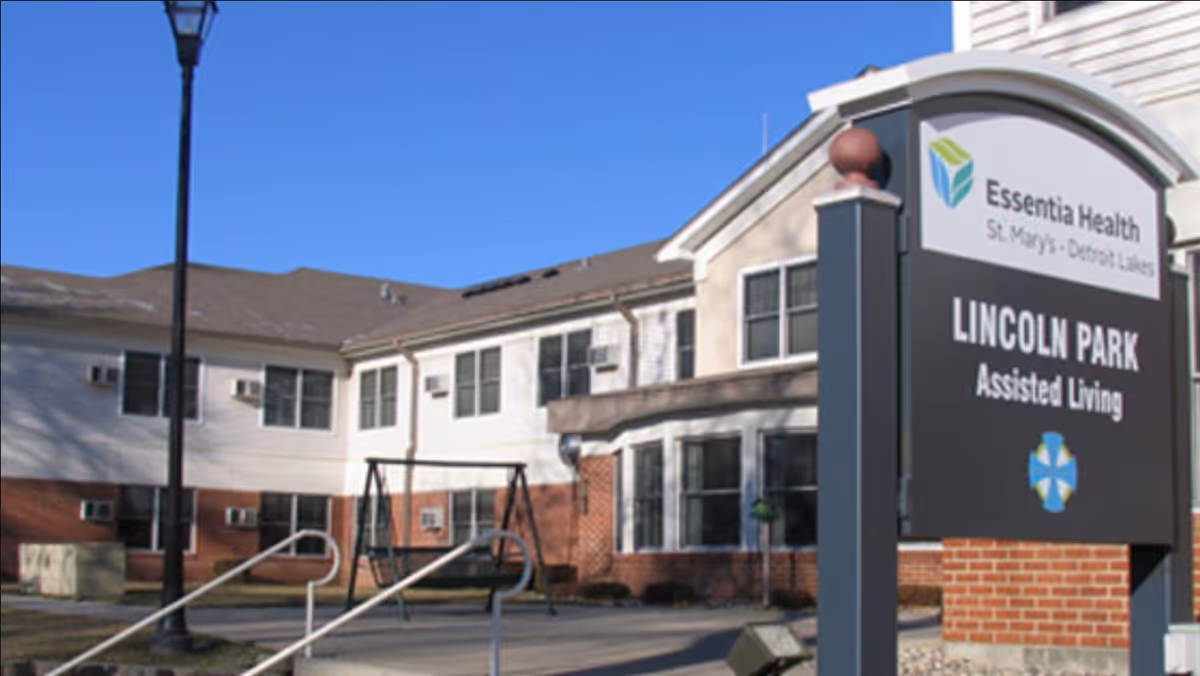 Exterior view of Essentia Health Lincoln Park Assisted Living facility showing a two-story building with white and brick walls, multiple windows, a swing set, a street lamp, and a large sign with the facility's name under a clear blue sky.
