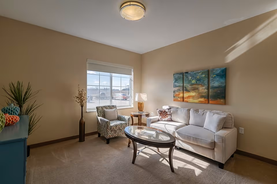 A cozy living room with a beige sofa, a patterned armchair, a glass-top coffee table, a side table with a lamp, a large window letting in natural light, and a three-panel abstract painting on the wall.