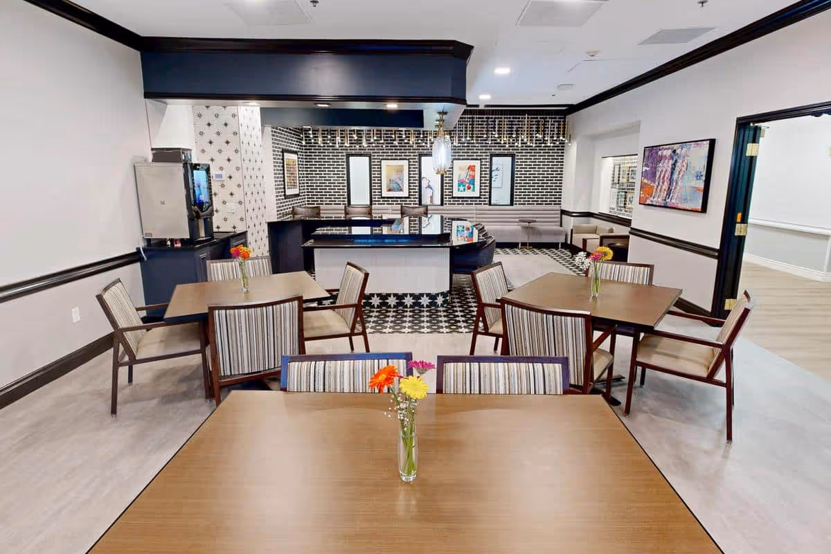 A dining area in Pegasus Landing of Tanglewood featuring several wooden tables with striped cushioned chairs. Each table has a small vase with colorful flowers. The back wall has a black and white patterned design with framed artwork and a counter area with bar stools. The room has light-colored flooring and white walls with dark trim.