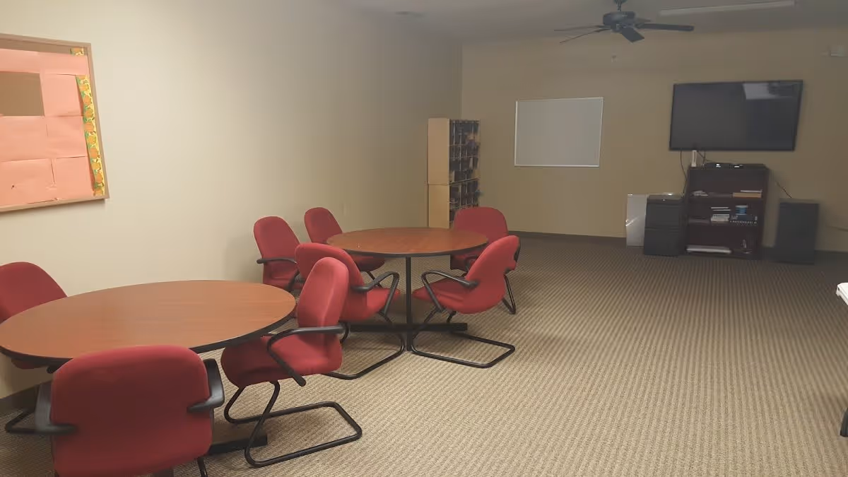 A room with beige walls and carpeted floor featuring two round wooden tables surrounded by red cushioned chairs. A bulletin board is mounted on the left wall, and a flat-screen TV is mounted on the far wall above a small shelving unit with electronic devices and books. A ceiling fan is visible on the ceiling.