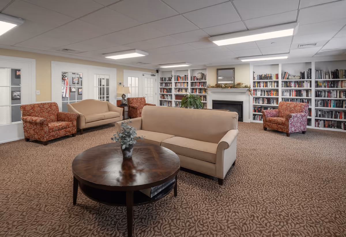 A cozy living room area with beige and patterned armchairs and sofas arranged around a round wooden coffee table with a small plant on it. The room features built-in bookshelves filled with books, a fireplace with a mirror above it, and soft overhead lighting. The carpet has a patterned design, and there are glass-paneled doors along one wall.