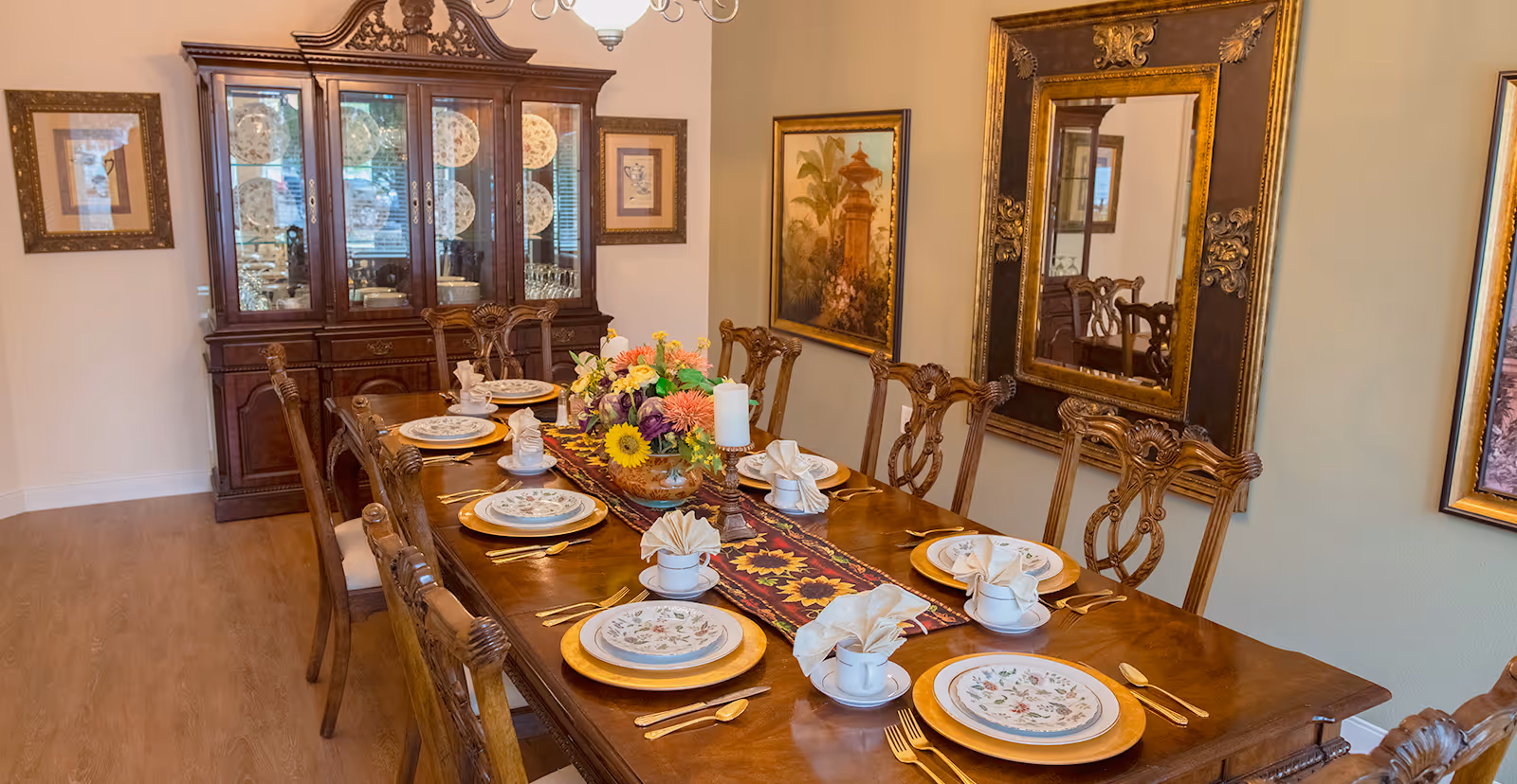 A formal dining room with a long wooden table set for eight, a floral centerpiece, china cabinet and ornate wall mirror.