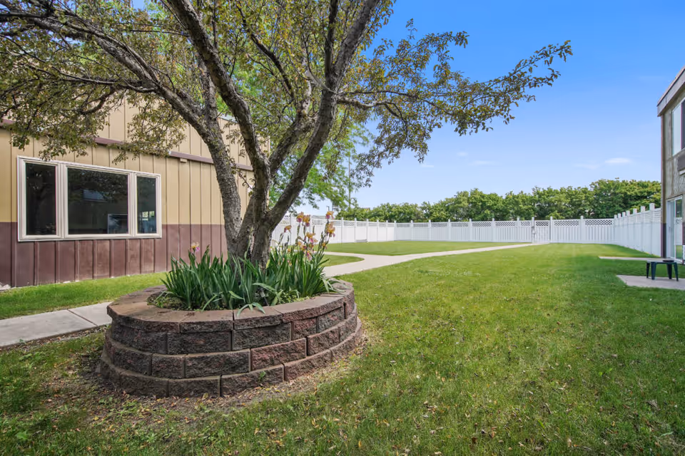 Courtyard with a tree in a circular brick planter, grassy lawn, winding walkway, surrounding buildings, and a white fence.