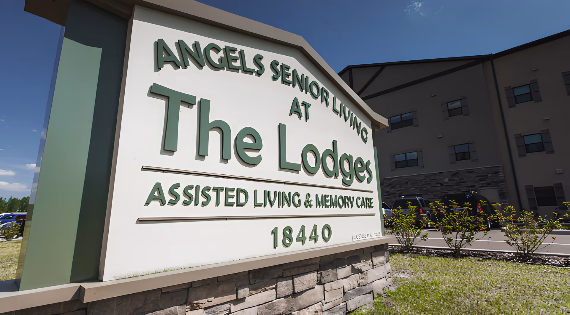 Outdoor view of a large sign for Angels Senior Living at The Lodges, an assisted living and memory care facility, with part of the building and parked cars visible in the background under a clear blue sky.