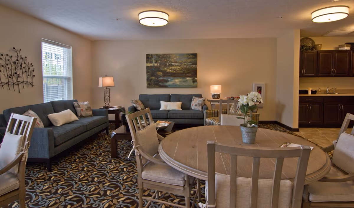 A cozy living room area with two blue sofas, a wooden round table with cushioned chairs, a small coffee table, and a kitchen area with dark wooden cabinets in the background. The room is decorated with a landscape painting, a metal wall art piece, table lamps, and a potted plant on the table. The floor has a patterned carpet.