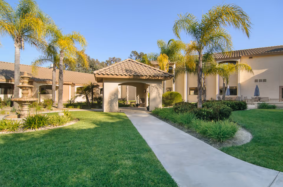 Outdoor courtyard area at Trabuco Hills Post-Acute with a paved walkway leading to a covered seating area. The courtyard is surrounded by green grass, palm trees, shrubs, and a multi-tiered stone fountain. The building has beige walls and a tiled roof under a clear blue sky.