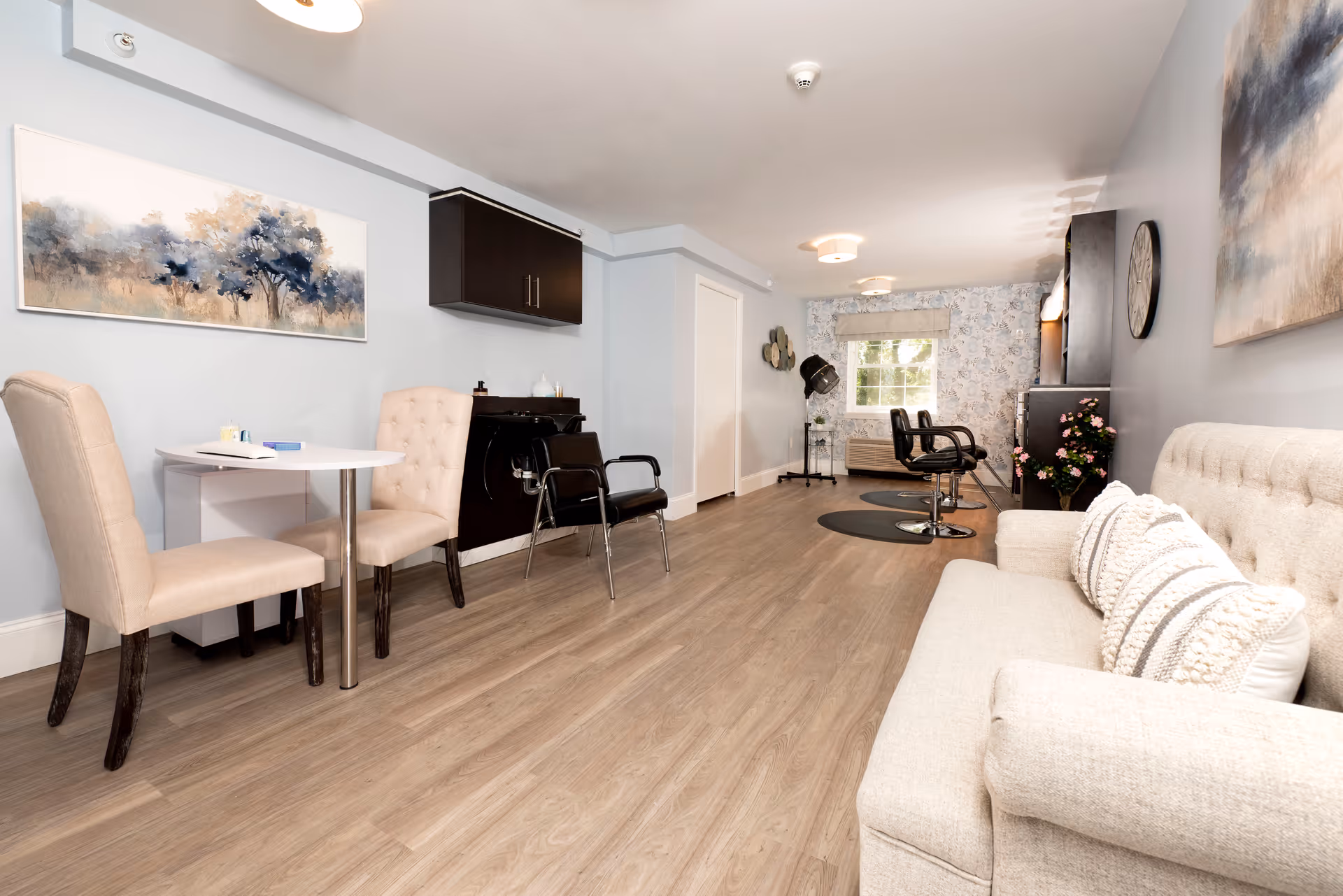 A bright and spacious room with light wood flooring and soft gray walls. On the right side, there is a beige upholstered sofa with decorative pillows and a large abstract painting above it. On the left side, there is a small white table with two beige upholstered chairs. Further back, there are two black salon chairs on circular mats in front of a window with floral curtains. The room has modern ceiling lights and a wall-mounted cabinet above a small counter area.